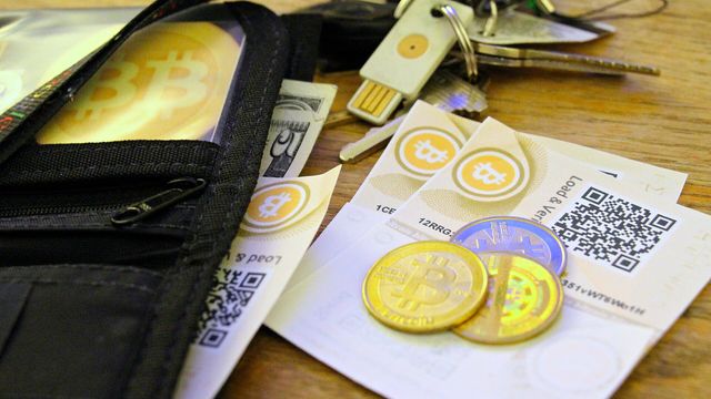 A hardware wallet on a keyring beside paper bitcoin wallets with QR codes, physical bitcoin coins and banknotes — FinCEN withdrew its unhosted-wallet and crypto-mixing reporting proposals